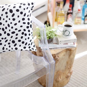 May include: A clear acrylic chair with a white pillow featuring black spots. A small wooden side table holds a coffee mug with a black initial, books, and a small plant. The scene suggests a modern, stylish interior.