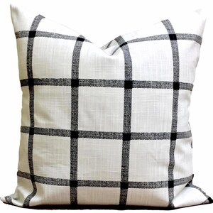 May include: Square white cushion featuring a black grid design. The cushion has a textured surface and a classic, geometric pattern. The black lines intersect to create squares, with solid black squares at the intersections.