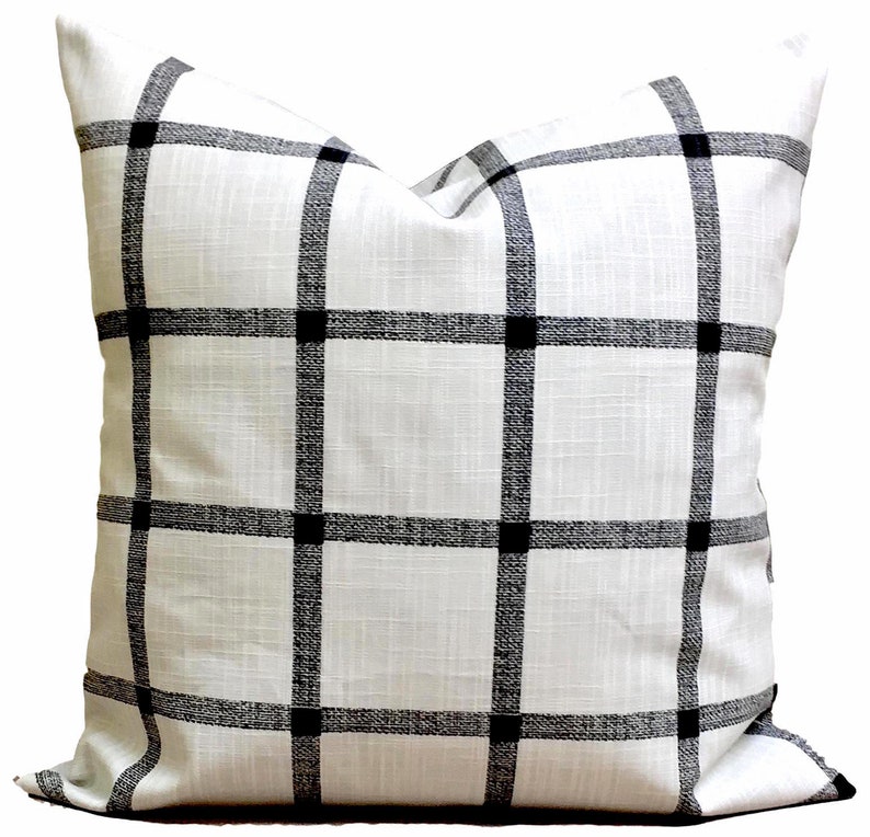 May include: Square white cushion featuring a black grid design. The cushion has a textured surface and a classic, geometric pattern. The black lines intersect to create squares, with solid black squares at the intersections.