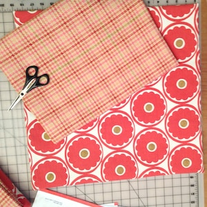 May include: Overhead shot of fabric samples and scissors. One fabric has a red and pink plaid pattern, and the other has a repeating floral design in red and gold on a white background. Black scissors are on the cutting mat.