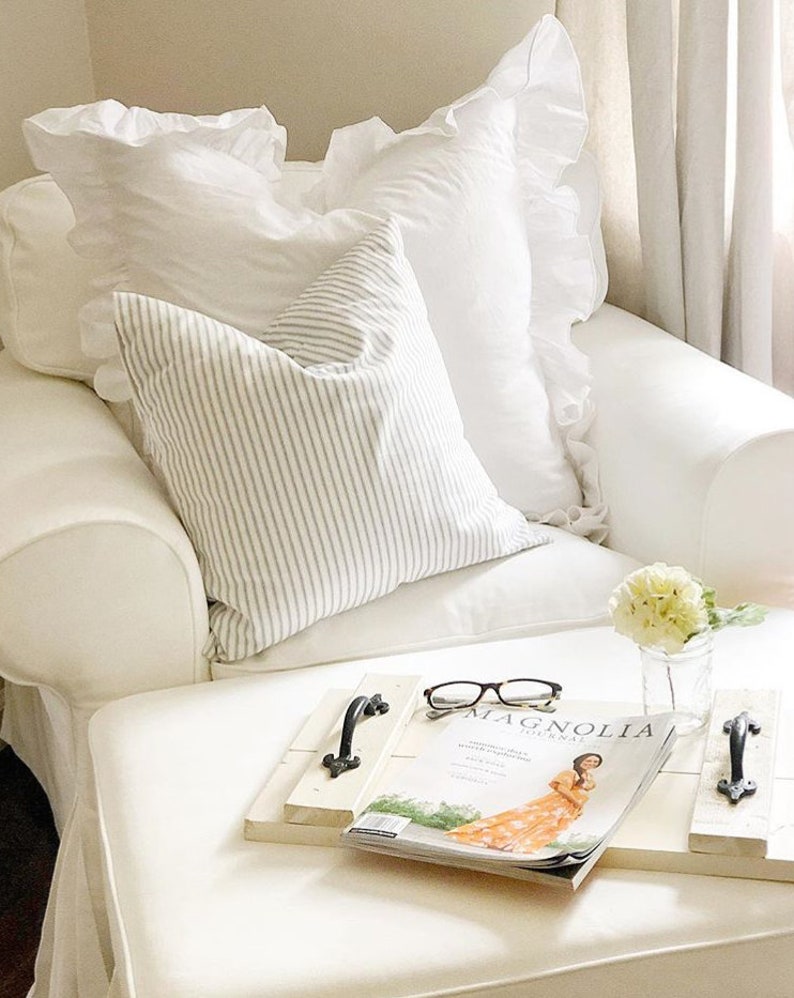 May include: A comfortable living room setting with a white armchair, white ruffled cushions, and a striped pillow. A wooden tray holds a magazine, spectacles, and a small vase with flowers, creating a relaxing atmosphere.