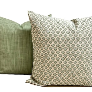 May include: Two square throw pillows. One pillow features a solid sage green fabric with a subtle cross-hatched texture. The second pillow displays a repeating pattern of small, sage green heart-shaped motifs on a light cream-colored background.