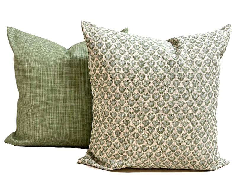 May include: Two square throw pillows. One pillow features a solid sage green fabric with a subtle cross-hatched texture. The second pillow displays a repeating pattern of small, sage green heart-shaped motifs on a light cream-colored background.