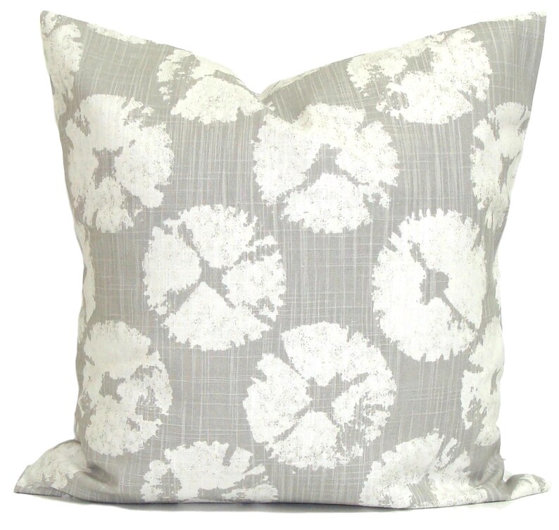 May include: A square cushion featuring a light grey backdrop with a repeating pattern of large, white, abstract floral shapes. The cushion has a soft, textured appearance, suitable for home decor.