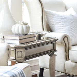 Puede incluir: Una calabaza de cerámica blanca se encuentra sobre dos libros en una mesa auxiliar plateada, junto a un sillón blanco. Una lámpara blanca y un cuenco decorativo también están sobre la mesa. El texto "HANDCRAFTED WITH ElemenOPillows" está en la parte inferior.