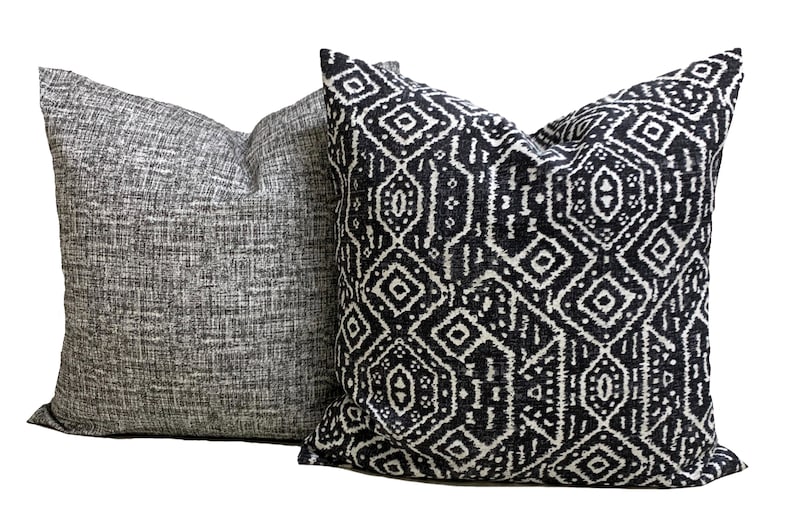 May include: Two square throw cushions. One is grey with a textured weave, the other has a black and white geometric design. The cushions are made of a soft fabric, suitable for a sofa or bed.