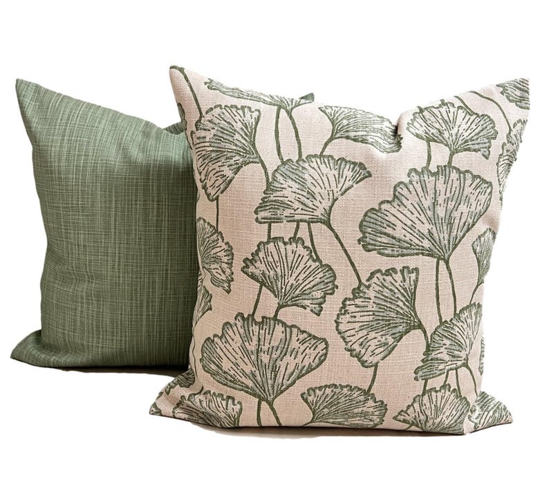 May include: Two square throw pillows. One pillow features a solid sage green fabric with a subtle cross-hatch texture. The second pillow displays a cream-colored background with a repeating pattern of dark green ginkgo leaf illustrations.