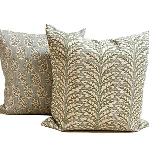 May include: Two square throw pillows featuring a light blue-grey fabric base. The front pillow displays a repeating vertical pattern of dark green fern-like leaves, while the back pillow features a repeating motif of small, dark green floral shapes.