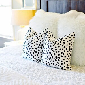 May include: Two white throw pillows with a black spot pattern rest on a white bedspread. A fluffy white pillow and a dark wooden headboard are in the background. A lit lamp is visible in the distance.