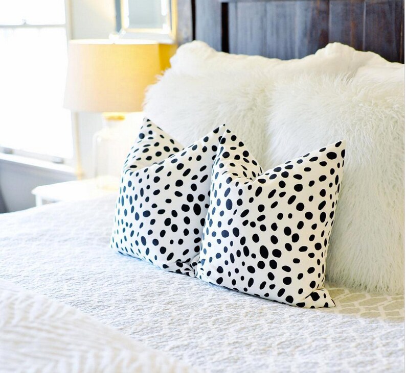 May include: Two white throw pillows with a black spot pattern rest on a white bedspread. A fluffy white pillow and a dark wooden headboard are in the background. A lit lamp is visible in the distance.