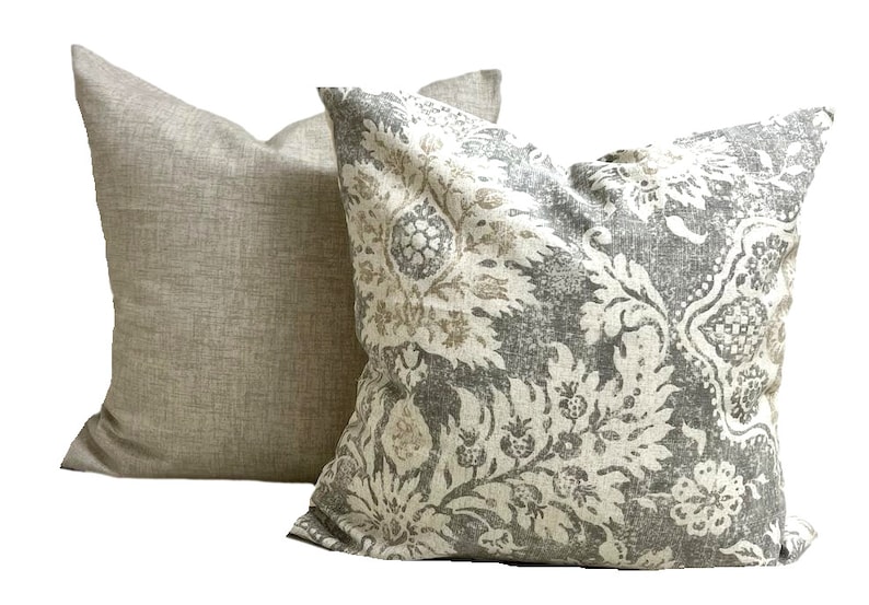 May include: Two decorative throw pillows. One pillow has a solid light tan fabric. The other pillow has a gray and off-white floral pattern. The pillows are square and appear to be made of a textured fabric.