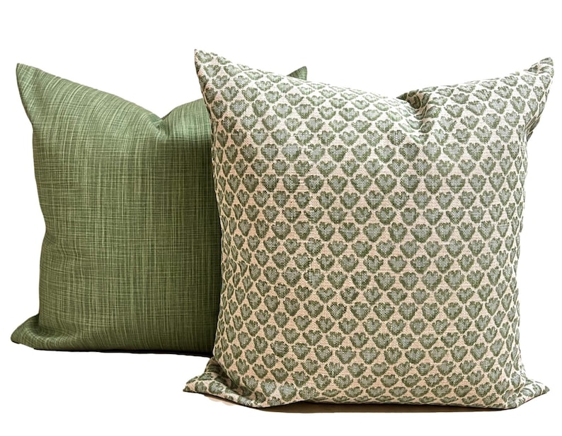 May include: Two square throw pillows. The pillow on the left is a solid sage green with a subtle cross-hatch texture. The pillow on the right features a repeating pattern of small, sage green heart-shaped motifs on a light cream-colored background.