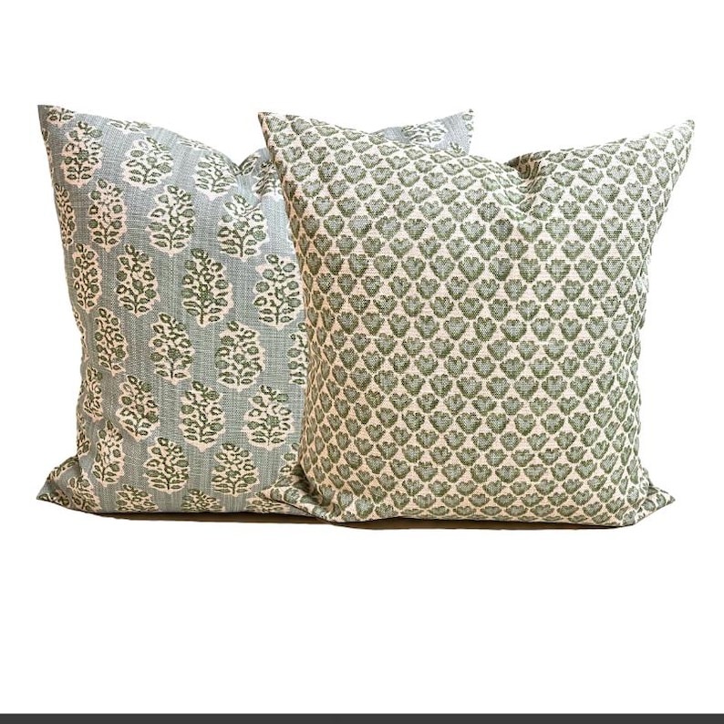 May include: Two square throw pillows featuring textured fabric. The left pillow displays a light blue background with a repeating green botanical leaf pattern. The right pillow features a cream background with a dense, repeating green heart pattern.