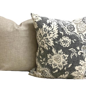 May include: Two decorative throw pillows. One is a solid light beige color with a textured appearance. The other pillow has a dark gray background with a large floral pattern in white and beige. Home decor.
