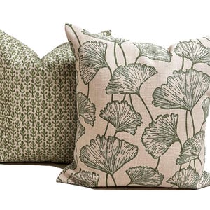 May include: Two square throw pillows with a textured beige fabric base. The front pillow features a large-scale green ginkgo leaf pattern, while the back pillow displays a smaller, repeating green botanical print of vertical leaf stems.
