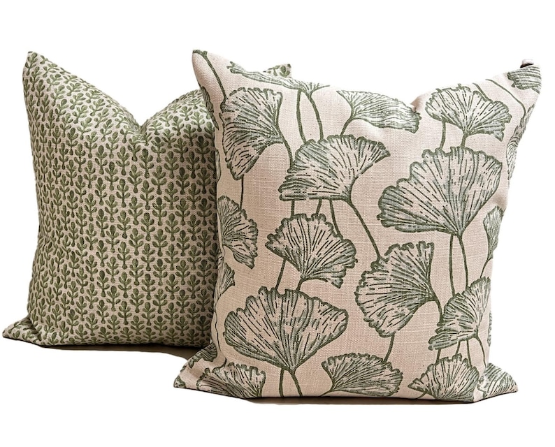 May include: Two square throw pillows with a textured beige fabric base. The front pillow features a large-scale green ginkgo leaf pattern, while the back pillow displays a smaller, repeating green botanical print of vertical leaf stems.