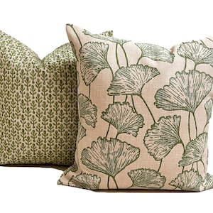 May include: Two square throw pillows with a textured beige fabric base. The front pillow features a large green ginkgo leaf pattern, while the back pillow displays a smaller, repeating green botanical print across its entire surface.