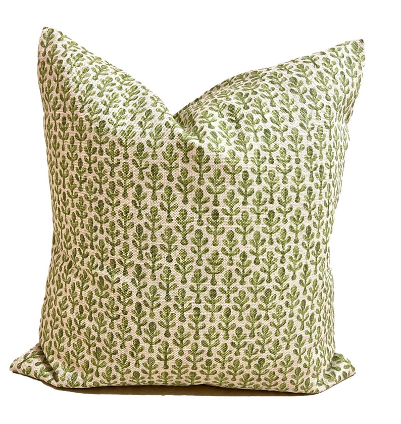 May include: A square throw pillow featuring a repeating pattern of small, stylized green leaf motifs printed on a textured, off-white fabric base. The pillow has a plump, filled appearance with visible seams along the edges and a woven textile finish.