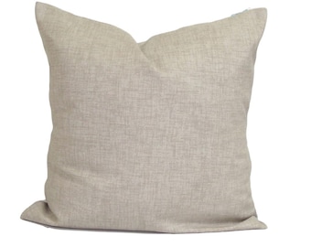 Pode incluir: Uma almofada quadrada de cor bege claro sólida com uma textura subtil que lembra um tecido. A almofada tem uma aparência macia e ligeiramente cheia com bordas limpas e retas, e um design simples e uniforme em toda a superfície frontal.
