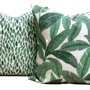 May include: Two decorative throw pillows. One pillow features a pattern of green leaf shapes on a white background. The other pillow has a tropical leaf design in shades of green and white. Both pillows are square.