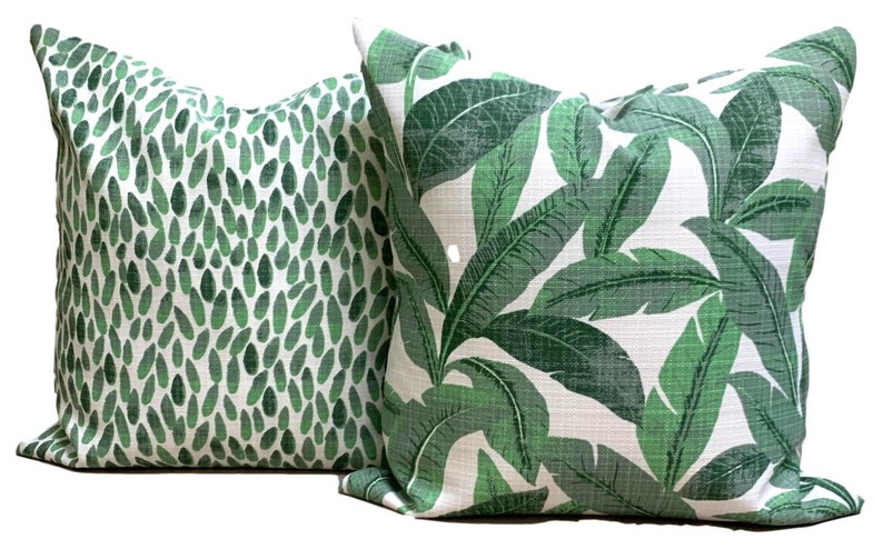 May include: Two decorative throw pillows. One pillow features a pattern of green leaf shapes on a white background. The other pillow has a tropical leaf design in shades of green and white. Both pillows are square.