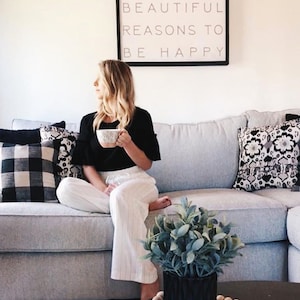 May include: A living room scene with a gray sectional sofa, decorative pillows, and a framed sign that reads "SO MANY BEAUTIFUL REASONS TO BE HAPPY." A person is seated on the sofa, holding a mug. A small plant in a black pot sits on a coffee table.