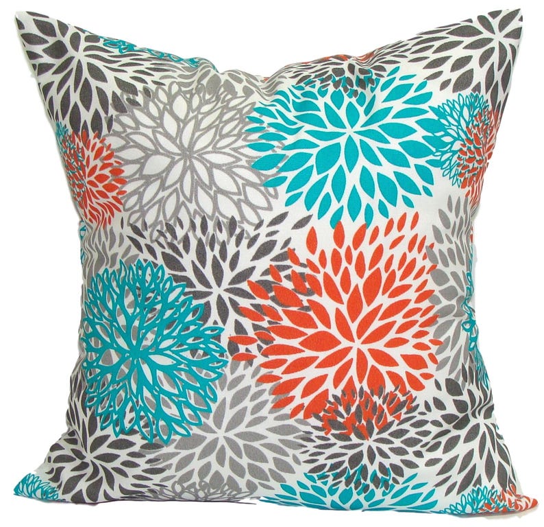 May include: Square throw pillow with a white background and a floral pattern. The flowers are in shades of gray, teal, and orange. The pillow is a decorative accent for a sofa or bed.