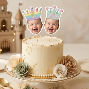 K&ouml;nnte beinhalten: Eine wei&szlig; glasierte Torte auf einem Tortenst&auml;nder, verziert mit Blattgold und Perlenr&auml;ndern. Zwei Tortenstecker zeigen l&auml;chelnde Babygesichter mit pastellfarbenen Kronen und der Zahl 1, umgeben von Papierblumen.