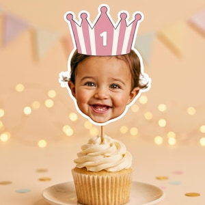 K&ouml;nnte beinhalten: Ein Cupcake-Topper mit einem Ausschnitt eines l&auml;chelnden Kindergesichts, das eine rosa-wei&szlig; gestreifte Krone mit der aufgedruckten Zahl 1 tr&auml;gt. Der Ausschnitt ist an einem d&uuml;nnen Holzstab befestigt, der in einem Cupcake steckt.