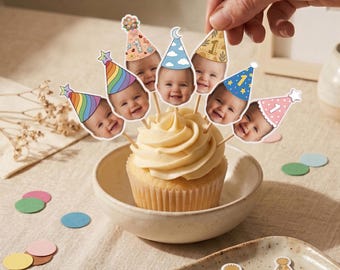 Könnte beinhalten: Ein Cupcake in einer Keramikschale, verziert mit mehreren Papier-Toppern, die das Gesicht eines Babys mit verschiedenen bunten Partyhüten und der aufgedruckten Zahl Eins zeigen. Weitere lose Partyhut-Topper und Holzspieße liegen daneben.