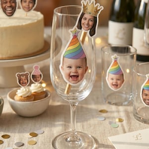 K&ouml;nnte beinhalten: Ein klares Weinglas mit einem individuellen Ausschnitt eines l&auml;chelnden Babys, das einen regenbogenfarbenen Partyhut tr&auml;gt. Ein goldfarbener Strohhalm steckt im Glas, verziert mit einem Ausschnitt einer Person mit einer goldenen Krone.