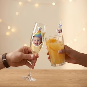 K&ouml;nnte beinhalten: Ein klares Sektglas mit einem Aufkleber eines l&auml;chelnden Kindes mit regenbogenfarbenem Partyhut sowie ein Trinkglas mit einem Strohhalm, der mit einem kleinen Ausschnitt eines Kindes mit pastellfarbenem Partyhut verziert ist.