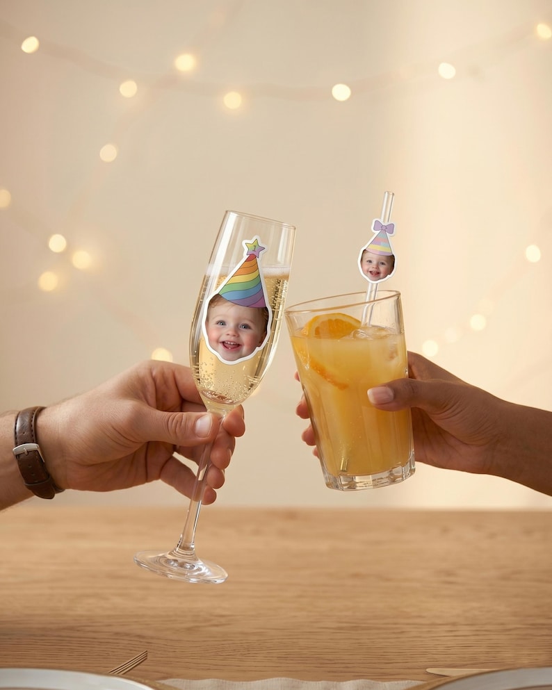 K&ouml;nnte beinhalten: Ein klares Sektglas mit einem Aufkleber eines l&auml;chelnden Kindes mit regenbogenfarbenem Partyhut sowie ein Trinkglas mit einem Strohhalm, der mit einem kleinen Ausschnitt eines Kindes mit pastellfarbenem Partyhut verziert ist.