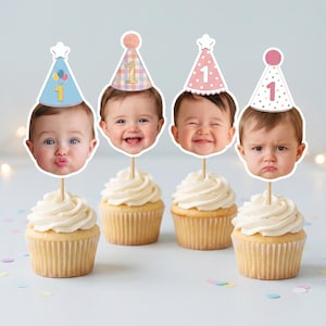 May include: Four cupcakes topped with custom photo face cutouts on wooden sticks. Each cutout features a baby wearing a different party hat with the number 1 printed on it. The hats include blue with balloons, plaid, pink with white dots, and white with dots.