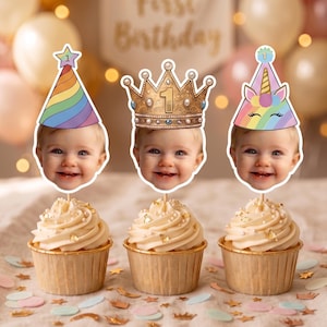 May include: Three cupcakes with white frosting and gold flakes, each topped with a custom photo cutout of a baby's face. The cutouts feature a rainbow party hat, a gold crown with the number 1, and a unicorn party hat with a horn.