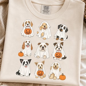 May include: A beige t-shirt featuring a graphic print of nine different dog breeds wearing white ghost costumes for Halloween. Some dogs are holding orange jack-o'-lantern candy buckets, while others are sitting next to small orange pumpkins.