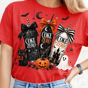 Könnte beinhalten: Ein rotes T-Shirt mit einer Grafik von drei Coke Zero Dosen, die mit Schleifen verziert sind, umgeben von Halloween-Elementen wie einer schwarzen Katze, einem Kürbis, einem Geist, Fledermäusen, Sternen und einer Mondsichel. Der Text Coke Zero ist zu sehen.