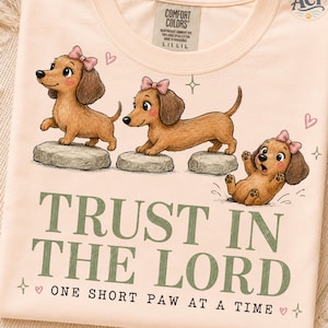 Może przedstawiać: Jasnobrzoskwiniowy t-shirt z trzema brązowymi jamnikami w różowych kokardkach. Dwa psy stoją na szarych kamieniach, a trzeci siedzi poniżej. Napis głosi: TRUST IN THE LORD ONE SHORT PAW AT A TIME. Przy szyi widoczna jest metka marki Comfort Colors.