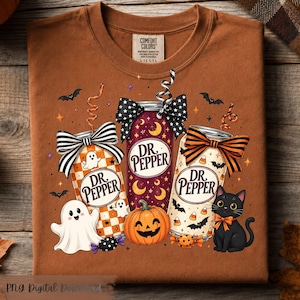 May include: A rust-colored t-shirt featuring a graphic of three Dr. Pepper cans decorated with bows, straws, and Halloween motifs including ghosts, a jack-o'-lantern, a black cat, bats, candy corn, and stars. The brand label reads Comfort Colors.