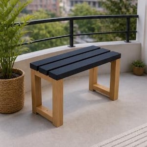 Puede incluir: Un pequeño banco para exteriores con asiento de listones negros y patas de madera marrón claro. El banco está colocado en un patio de hormigón junto a una planta en maceta. El banco mide aproximadamente 91 cm de largo.