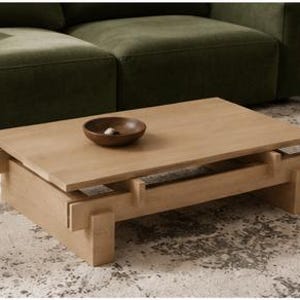 May include: A light-colored wooden coffee table with a rectangular top and a lower shelf. The table has a modern design with a bowl on top. The table is in front of a green sofa.