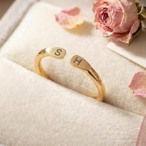 May include: A gold-toned open ring featuring the letters S and H engraved on the ends. The ring is positioned inside a light grey velvet jewellery box alongside two small, dried pink rosebuds and a delicate pink ribbon.