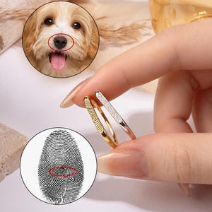 May include: Two thin metal rings, one gold-toned with a textured fingerprint pattern and one silver-toned with a delicate engraved design. The rings are displayed alongside circular insets showing a dog nose and a detailed human fingerprint.