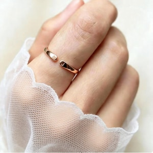 Puede incluir: Un anillo abierto de oro rosa con dos extremos redondeados, cada uno grabado con una letra. Un extremo muestra la letra A y el otro extremo muestra la letra J. El anillo tiene un acabado metálico pulido y un diseño de banda delgada.
