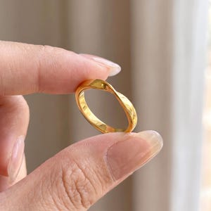 May include: A polished gold-tone ring featuring a smooth, continuous twisted band design. The metal surface has a reflective, high-shine finish that catches the light, creating a subtle undulating shape around the circumference of the circular band.