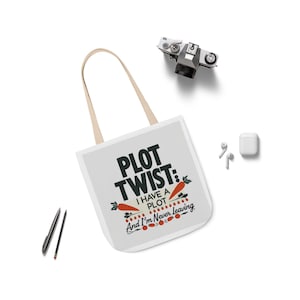 Puede incluir: Bolso tote blanco con el texto "PLOT TWIST: I HAVE A PLOT And I'm Never Leaving" en negro, rojo y naranja. El bolso tiene asas beige y está sobre una superficie blanca. También se ven una cámara, bolígrafos y auriculares.