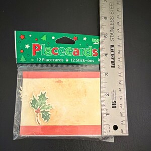 May include: A package of 12 Christmas placecards with 12 stick-ons, featuring a cream-colored card with red borders and a holly leaf graphic. A stainless steel ruler is placed alongside, showing the package is approximately 6 inches in height.