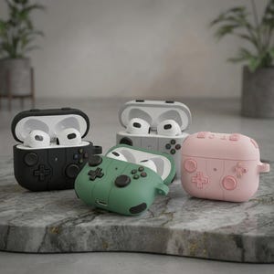 Puede incluir: Cuatro coloridas fundas para AirPod en negro, verde, gris y rosa, cada una diseñada para parecerse a un mando de videojuegos. Las fundas están abiertas, revelando los auriculares blancos en su interior. Las fundas están sobre una superficie de mármol.