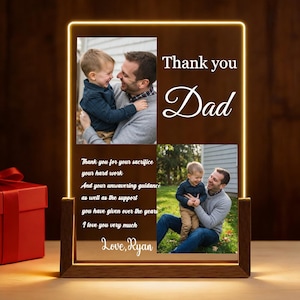 Puede incluir: Un marco de fotos LED personalizado con base de madera. El marco muestra dos fotos de un padre y un hijo, con las palabras "Thank you Dad" y un mensaje sincero. El marco está iluminado con una luz cálida y amarilla.