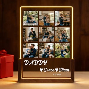 May include: A personalized wooden photo display with nine family photos. The display has a warm, glowing LED border and features the words "DADDY", "Grace", "Ethan", and the date "6.21.2026". A red gift box is visible in the background.